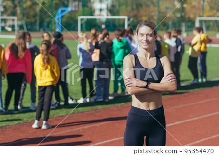 A female physical education teacher at school smiles and looks at the camera, an athlete trainer on the background of children, portrait. A female physical education teacher at school smiles and looks at the camera, an athlete trainer on the background of children, portrait. 95354470