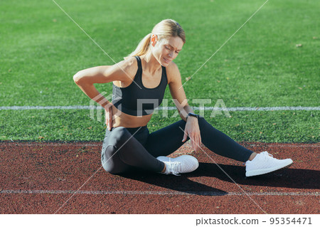 Injured female athlete sits on the stadium treadmill, has severe back pain, holds her hand by her side, massages a muscle. Injured female athlete sits on the stadium treadmill, has severe back pain, holds her hand by her side, massages a muscle. 95354471