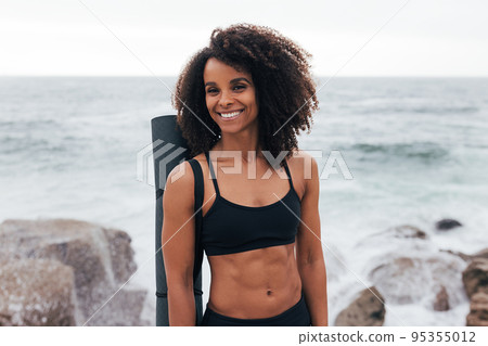 Portrait of a muscular woman with a mat standing by the ocean and looking away Portrait of a muscular woman with a mat standing by the ocean and looking away 95355012