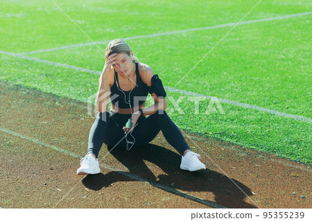 Tired sportswoman sitting on treadmill, blonde resting after jogging, woman listening to music and online radio podcast, motivational audio book in headphones. 95355239