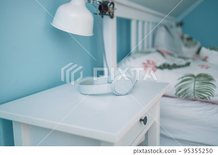 White wireless headphones lie on the bedside table near the bed in hotel room. White wood bedside table. White wireless headphones lie on the bedside table near the bed in hotel room. White wood bedside table. 95355250