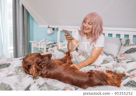 Beautiful young adult cheerful woman taking selfie on smartphone with her dog Irish Setter in morning in bed Beautiful young adult cheerful woman taking selfie on smartphone with her dog Irish Setter in morning in bed 95355252