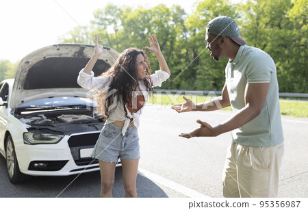 Millennial multiracial couple having conflict, shouting at each other on road near broken car Millennial multiracial couple having conflict, shouting at each other on road near broken car 95356987