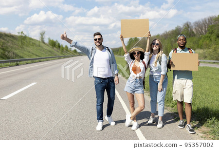 Happy diverse friends standing on highway with empty signs, stopping car, going on trip together, outdoors 95357002