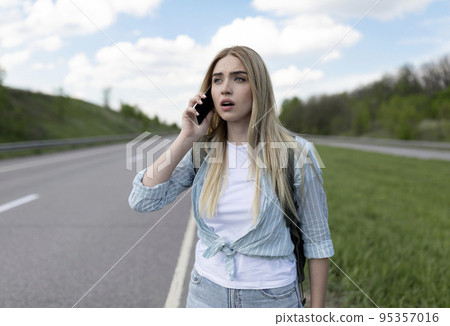 Confused young blonde woman standing along road, feeling lost, calling on cellphone, asking for help, free space 95357016
