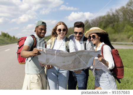 Group of multinational young friends looking at map, choosing route during road trip, hitchhiking on highway 95357083