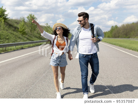 Handsome young guy walking along road with his girlfriend, holding hands, hitchhiking for car, stopping transport 95357087
