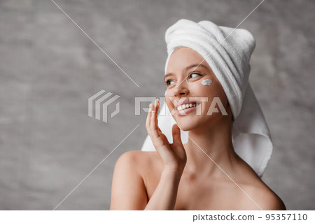 Female skincare concept. Lady applying moisturizer cream under eyes, standing in bathroom, looking aside and smiling 95357110