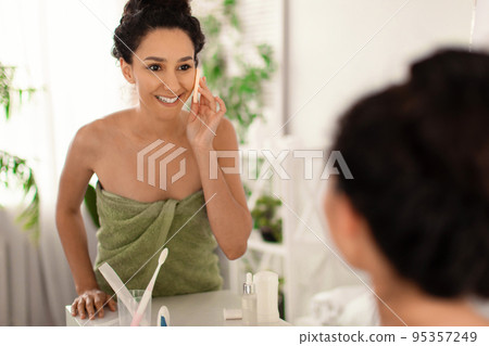 Happy millennial woman wearing towel, applying powder with cosmetic sponge near mirror at home, copy space Happy millennial woman wearing towel, applying powder with cosmetic sponge near mirror at home, copy space 95357249