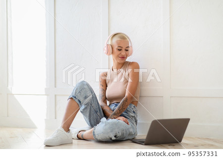 Happy young lady using laptop for online communication and wearing headphones, sitting on floor against light wall 95357331