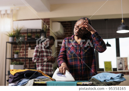 Tired modern african american wife ironing clothes while relaxed distracted husband uses cellphone. Disappointed and exhausted wife frustrated at taking care of all household chores alone without help 95358165