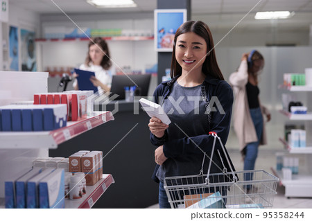 Woman client holding shopping basket full with health care products, choosing to buy vitamin supplements for for immune system. Pharmacy space with clients and pharmaceutical drugs 95358244