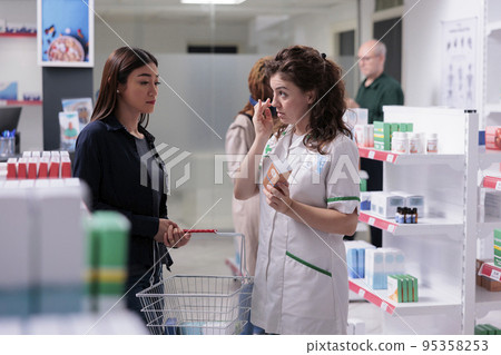 Woman customer talking with helpful pharmacy specialist about SPF prescription in drugstore. Pharmacist in medical uniform selling pharmaceutical product, explaining healthcare importance. Woman customer talking with helpful pharmacy specialist about SPF prescription in drugstore. Pharmacist in medical uniform selling pharmaceutical product, explaining healthcare importance. 95358253