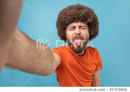 Portrait of childish silly man with Afro hairstyle wearing orange T-shirt taking selfie or has livestream, showing tongue out, point of view photo. Indoor studio shot isolated on blue background. 95359668