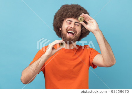 Portrait of funny laughing man with Afro hairstyle wearing orange T-shirt standing holding bitcoins, paying attention at new digital cryptocurrency. Indoor studio shot isolated on blue background. 95359670