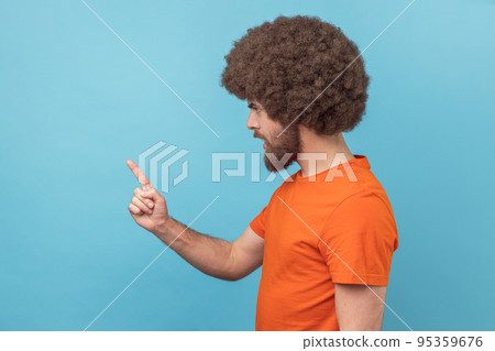 Side view of serious man with Afro hairstyle wearing orange T-shirt standing with admonishing gesture, scolding for mistake and warning. Indoor studio shot isolated on blue background. Side view of serious man with Afro hairstyle wearing orange T-shirt standing with admonishing gesture, scolding for mistake and warning. Indoor studio shot isolated on blue background. 95359676