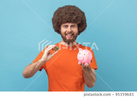 Portrait of man with Afro hairstyle wearing orange T-shirt holding piggy bank and golden bitcoin, looking at camera with happy expression. Indoor studio shot isolated on blue background. 95359683