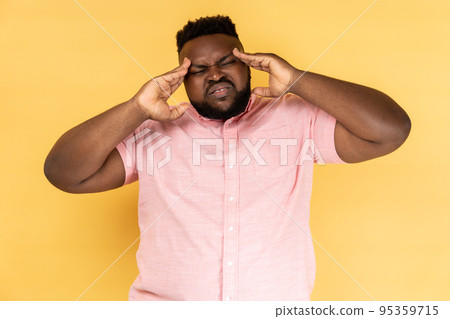 Portrait of unhealthy sick young adult man wearing pink shirt massaging temples, suffering from headache, migraine, frowning his face. Indoor studio shot isolated on yellow background. 95359715