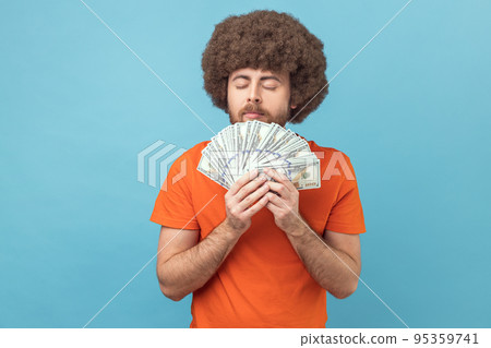 Portrait of man with Afro hairstyle wearing orange T-shirt smelling dollar banknotes with pleasure expression, enjoying big money profit and rich life. Indoor studio shot isolated on blue background. Portrait of man with Afro hairstyle wearing orange T-shirt smelling dollar banknotes with pleasure expression, enjoying big money profit and rich life. Indoor studio shot isolated on blue background. 95359741