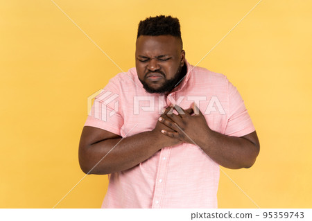 Cardiac problems. Portrait of young adult fatigued man wearing pink shirt clasping chest, feeling acute pain, heart attack from anxiety, infarction. Indoor studio shot isolated on yellow background. Cardiac problems. Portrait of young adult fatigued man wearing pink shirt clasping chest, feeling acute pain, heart attack from anxiety, infarction. Indoor studio shot isolated on yellow background. 95359743