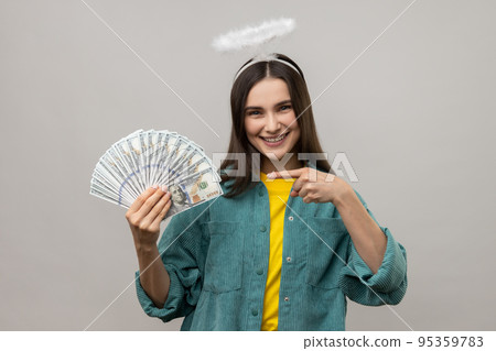 Cheerful dark haired woman with halo on head pointing finger on bunch of dollar banknotes and smiling, profit, wearing casual style jacket. Indoor studio shot isolated on gray background. Cheerful dark haired woman with halo on head pointing finger on bunch of dollar banknotes and smiling, profit, wearing casual style jacket. Indoor studio shot isolated on gray background. 95359783