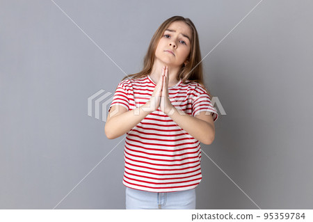Please help me or forgive. Portrait of worry sad little girl wearing striped T-shirt standing with palm hands and looking at camera pleased pleading. Indoor studio shot isolated on gray background. 95359784
