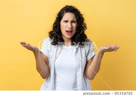 Portrait of unhappy stressed beautiful woman with dark wavy hair standing raised arms and asking, expressing negative emotions. Indoor studio shot isolated on yellow background. 95359785