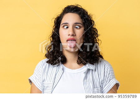 Portrait of woman with dark wavy hair standing with crossed eyes and tongue out, having foolish facial expression, childish behavior. Indoor studio shot isolated on yellow background. Portrait of woman with dark wavy hair standing with crossed eyes and tongue out, having foolish facial expression, childish behavior. Indoor studio shot isolated on yellow background. 95359789