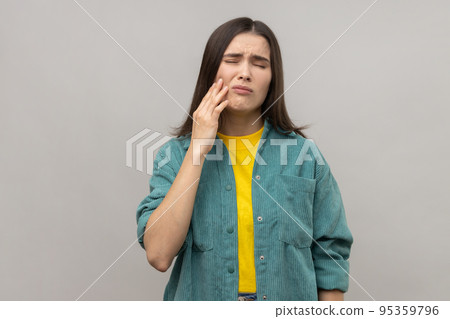 Unhealthy woman touching cheek, closing eyes with expression of terrible suffer from toothache, sensitive teeth, cavities, wearing casual style jacket. Indoor studio shot isolated on gray background. Unhealthy woman touching cheek, closing eyes with expression of terrible suffer from toothache, sensitive teeth, cavities, wearing casual style jacket. Indoor studio shot isolated on gray background. 95359796