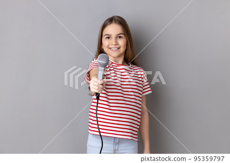 Portrait of cute little girl wearing striped T-shirt posing with microphone in hands, offers mic, asking questions, looking at camera. Indoor studio shot isolated on gray background. Portrait of cute little girl wearing striped T-shirt posing with microphone in hands, offers mic, asking questions, looking at camera. Indoor studio shot isolated on gray background. 95359797