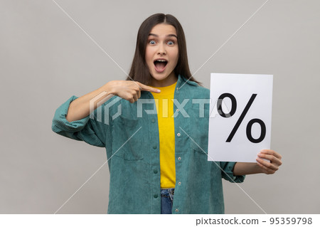 Amazed surprised woman holding and pointing at paper with percent sign inscription, looking at camera with open mouth, wearing casual style jacket. Indoor studio shot isolated on gray background. Amazed surprised woman holding and pointing at paper with percent sign inscription, looking at camera with open mouth, wearing casual style jacket. Indoor studio shot isolated on gray background. 95359798