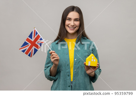 Portrait of smiling holding Great Britain flag and paper house, dreaming to buy accommodation in England, wearing casual style jacket. Indoor studio shot isolated on gray background. Portrait of smiling holding Great Britain flag and paper house, dreaming to buy accommodation in England, wearing casual style jacket. Indoor studio shot isolated on gray background. 95359799