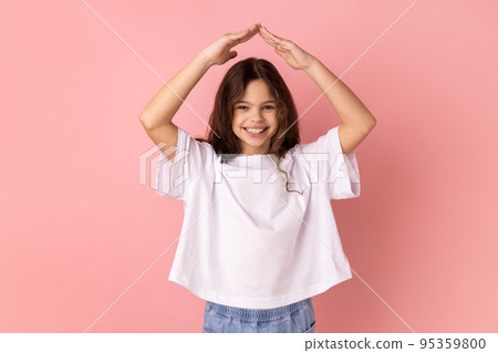 Portrait of little girl wearing white T-shirt with charming smile holding hands in gesture of house roof over head and smiling, feeling safe. Indoor studio shot isolated on pink background. Portrait of little girl wearing white T-shirt with charming smile holding hands in gesture of house roof over head and smiling, feeling safe. Indoor studio shot isolated on pink background. 95359800