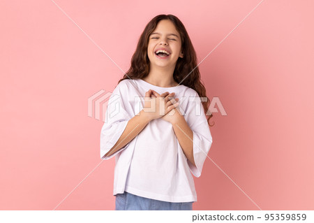 Portrait of crazy optimistic little girl wearing white T-shirt hearing funny joke, laughing out loud, holding belly, expressing positive emotions. Indoor studio shot isolated on pink background. 95359859