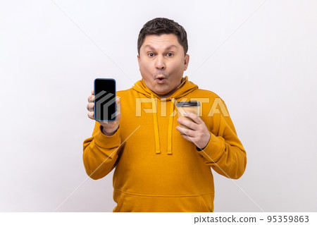 Portrait of astonished surprised man of middle age holding smart phone with blank screen and disposable cup of coffee, wearing urban style hoodie. Indoor studio shot isolated on white background. Portrait of astonished surprised man of middle age holding smart phone with blank screen and disposable cup of coffee, wearing urban style hoodie. Indoor studio shot isolated on white background. 95359863