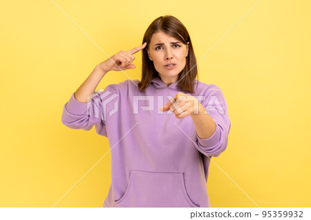 You are crazy. Attractive woman with hair holding finger near temple and pointing on you, showing stupid gesture, wearing purple hoodie. Indoor studio shot isolated on yellow background. You are crazy. Attractive woman with hair holding finger near temple and pointing on you, showing stupid gesture, wearing purple hoodie. Indoor studio shot isolated on yellow background. 95359932