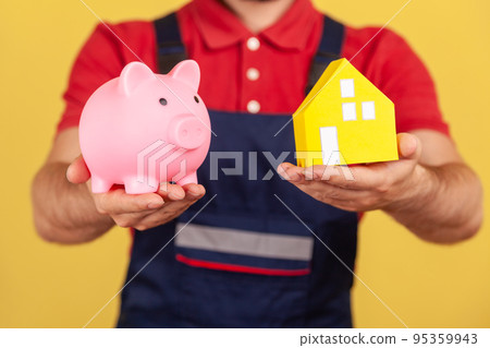 Portrait of unknown anonymous man wearing blue uniform and red T-shirt showing piggy bank and paper house, saving money buying real estate. Indoor studio shot isolated on yellow background. Portrait of unknown anonymous man wearing blue uniform and red T-shirt showing piggy bank and paper house, saving money buying real estate. Indoor studio shot isolated on yellow background. 95359943