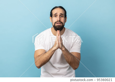 Please, I'm begging forgive. Portrait of upset worried man with beard wearing white T-shirt looking with imploring desperate grimace, praying for help. Indoor studio shot isolated on blue background. Please, I'm begging forgive. Portrait of upset worried man with beard wearing white T-shirt looking with imploring desperate grimace, praying for help. Indoor studio shot isolated on blue background. 95360021