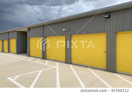 Storage unit buildings site outside, owners property. Road with markings leading to garages with yellow roller doors. 95360212