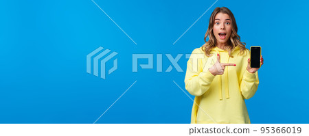 Impressed happy cheerful blond girl explain something as pointing mobile phone display, talking about new application or sharing her vacation photos, hold smartphone, look camera Impressed happy cheerful blond girl explain something as pointing mobile phone display, talking about new application or sharing her vacation photos, hold smartphone, look camera 95366019