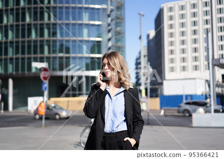Portrait of businesswoman in suit, standing on street with mobile phone, on call with someone, smiling while having telephone conversation 95366421