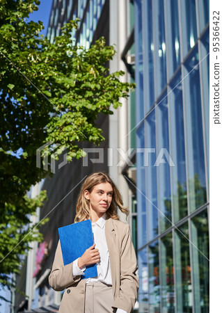 Portrait of confident saleswoman in her 30s, posing near office building on street, wearing beige suit, holding folder with documents 95366422