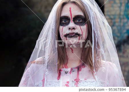 Portrait of zombie woman in wedding dress with veil and stage makeup looking into camera. Girl applied white makeup and blood stains on her face, touching face Portrait of zombie woman in wedding dress with veil and stage makeup looking into camera. Girl applied white makeup and blood stains on her face, touching face 95367894