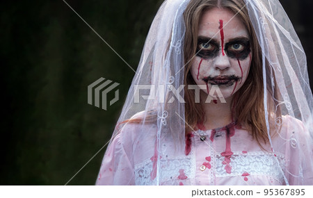 Portrait of zombie woman in wedding dress with veil and stage makeup looking into camera. Girl applied white makeup and blood stains on her face, touching face Portrait of zombie woman in wedding dress with veil and stage makeup looking into camera. Girl applied white makeup and blood stains on her face, touching face 95367895