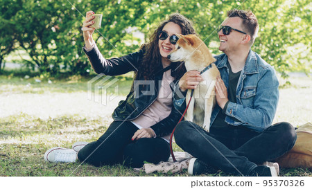 Happy couple is taking selfie with their dog using smartphone sitting on grass in the park, people are posing, talking and petting shiba inu puppy. Photograph and animals concept. Happy couple is taking selfie with their dog using smartphone sitting on grass in the park, people are posing, talking and petting shiba inu puppy. Photograph and animals concept. 95370326