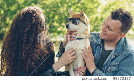 Cheerful young couple is having fun with dog putting sunglasses on its eyes and laughing while spending weekend in the park. Nature, modern lifestyle and animals concept. 95370327