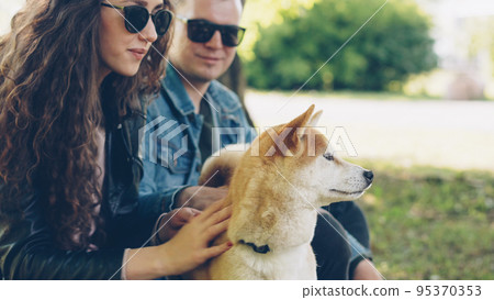 Happy couple girlfriend and boyfriend are patting cute puppy in the park and talking. Woman and man are wearing casual clothes and sunglasses, beautiful city park is in background. 95370353