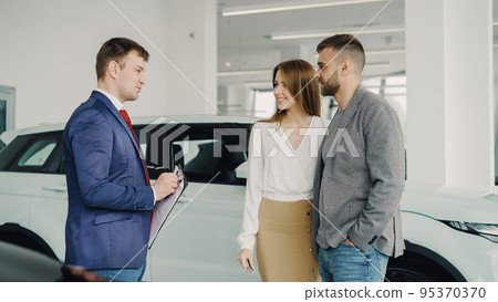 Car dealership worker is talking to beautiful couple young bearded man and his attractive wife while standing near luxury car in auto salon. Choosing new automobile concept. Car dealership worker is talking to beautiful couple young bearded man and his attractive wife while standing near luxury car in auto salon. Choosing new automobile concept. 95370370