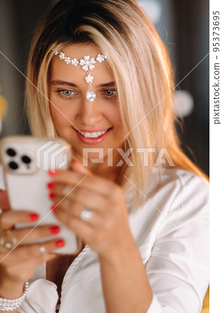 A positive young woman in a white dress and an ornament on her head was smiling, holding a phone in her hands and looking into it 95373695
