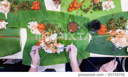 Overhead view of people savoring Indian spicy food served on banana leaf at restaurant. Known as banana leaf rice. 95376742